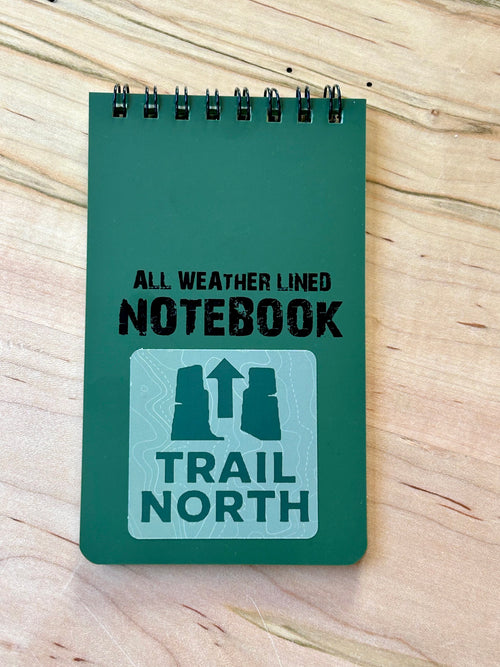 Green notebook with 'All Weather Lined Notebook Trail North' text on a wooden surface